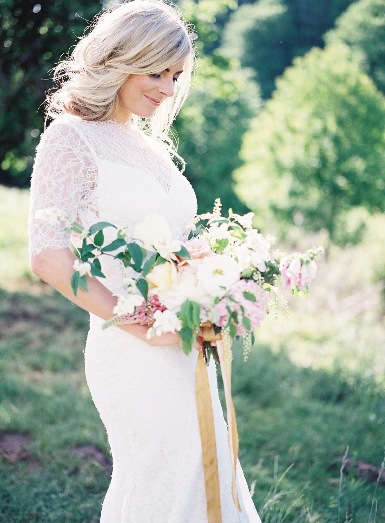 Backlit blonde bride in lace gown holding loose garden bouquet with gold ribbon in sunlit meadow