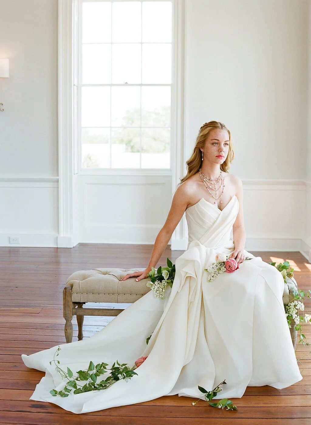Full-length editorial bride seated on tufted bench in sunlit white room, ivory gown with trailing greenery and pink rose