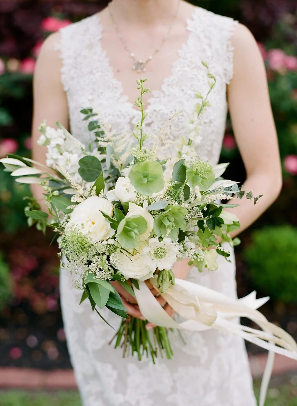 Bride in lace gown holding a loose white-and-green garden bouquet (ranunculus, hellebore, olive) against a rose garden