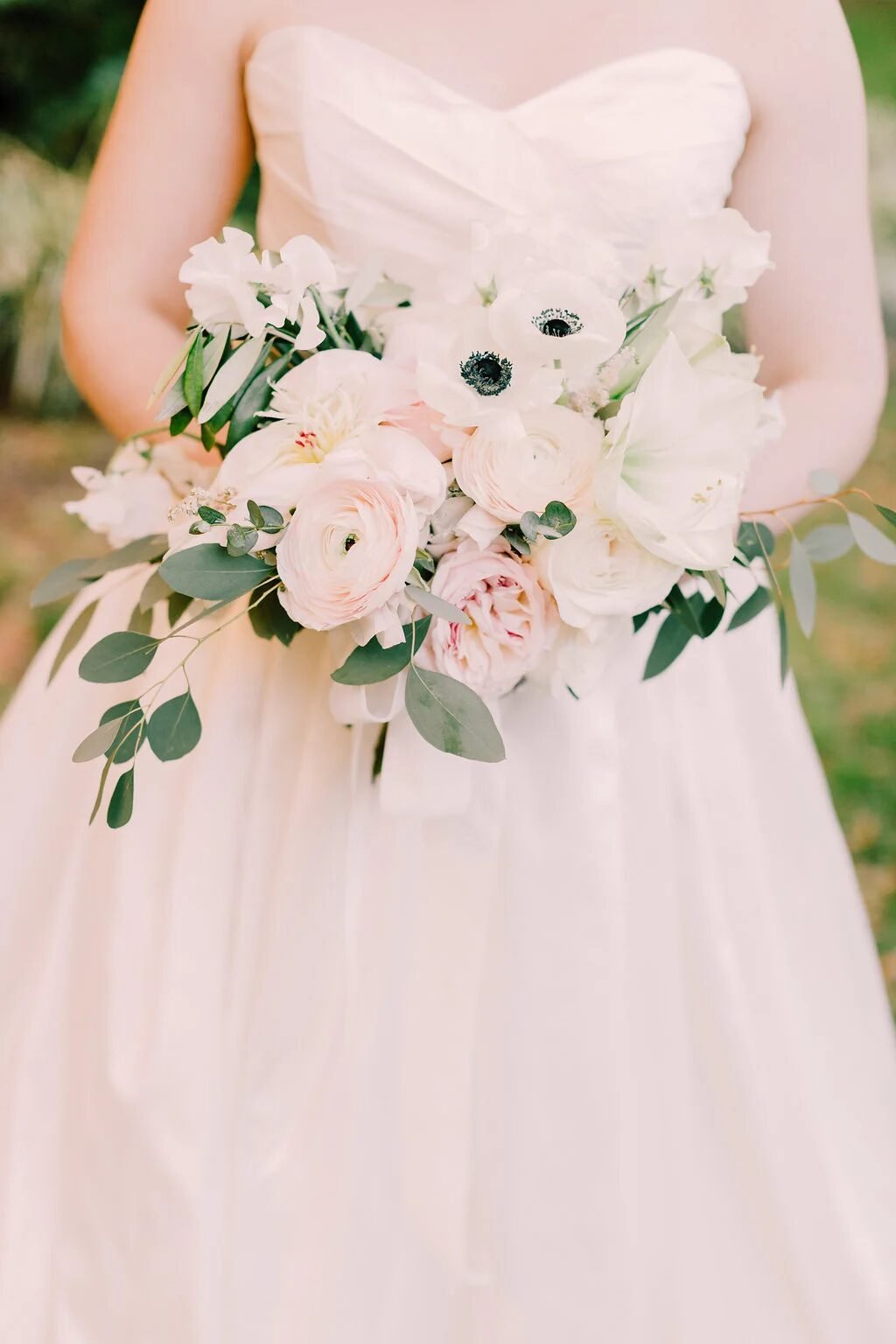 Bridal bouquet of white anemones, blush ranunculus, garden roses and eucalyptus against ivory gown