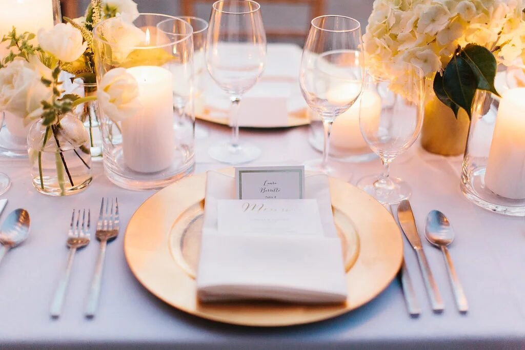 Candlelit gold-charger place setting with calligraphed menu, white blooms and warm taper glow