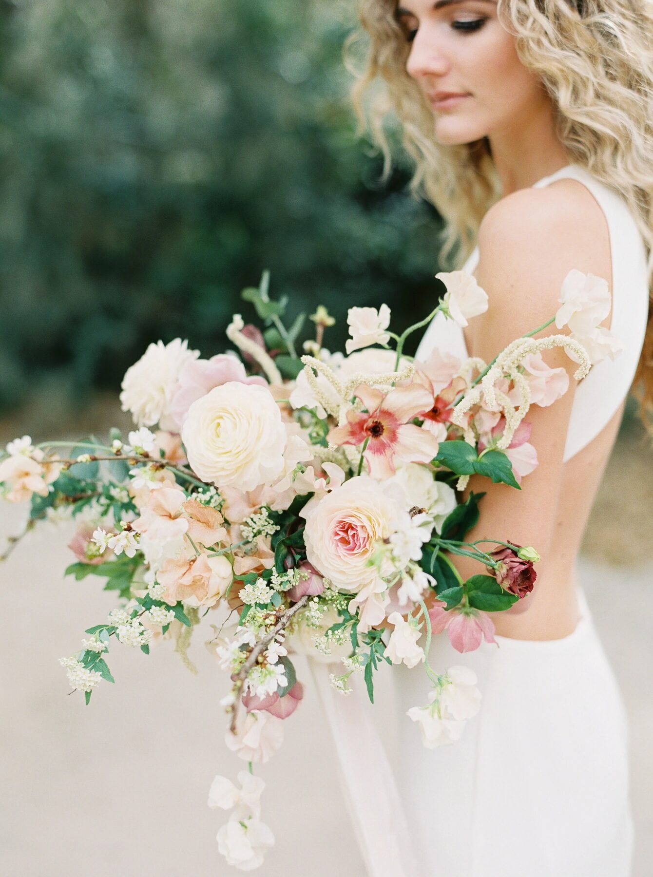 Bride in backless gown holding an airy cascading pastel bouquet of garden roses, sweet peas and hellebore, shot on film