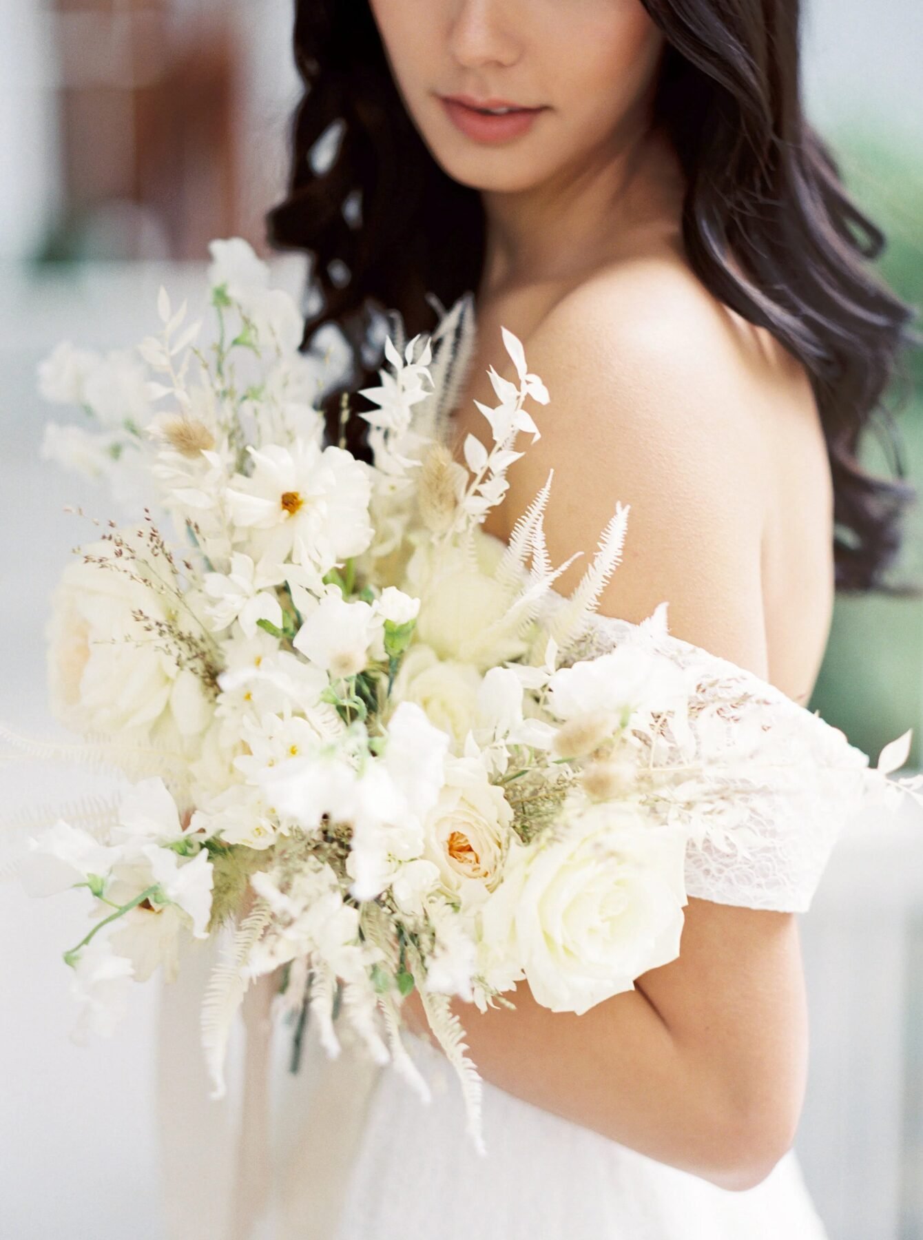 Tight detail of an all-white textured bridal bouquet (garden roses, cosmos, dried ferns, bunny tail) held against an off-shoulder lace gown
