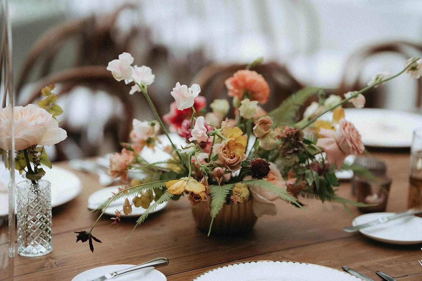Earthy autumn-toned centerpiece (terracotta, mustard, sage) on a wood table with shallow-focus place settings receding into creamy bokeh