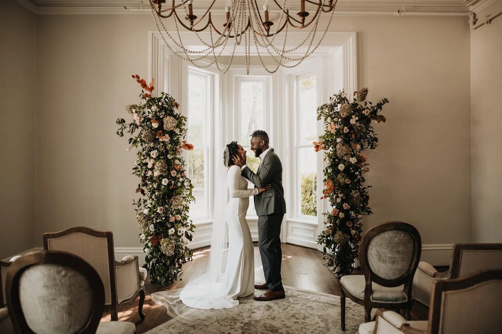 Intimate indoor elopement: couple embracing between two lush floral columns in a bright bay window, warm moody parlor with chandelier and vintage chairs