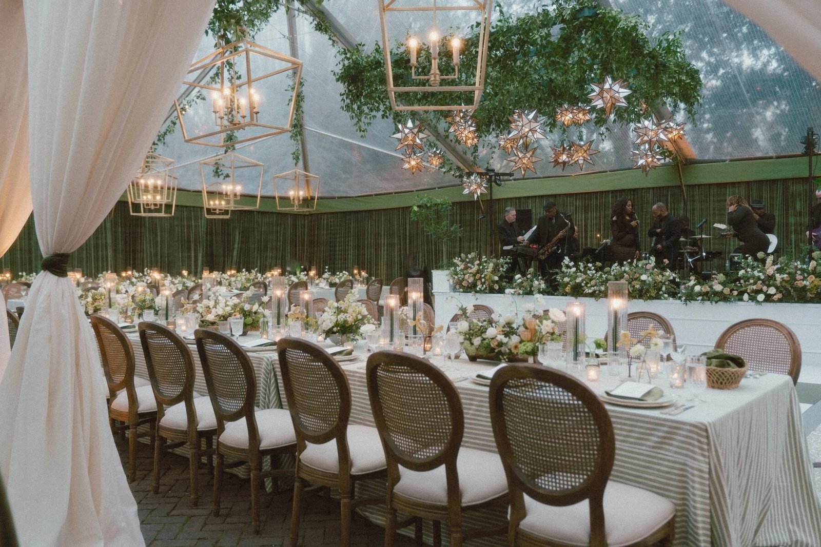Garden-style tented reception with olive bamboo backdrop, brass lanterns, trailing greenery overhead and candlelit dinner tables