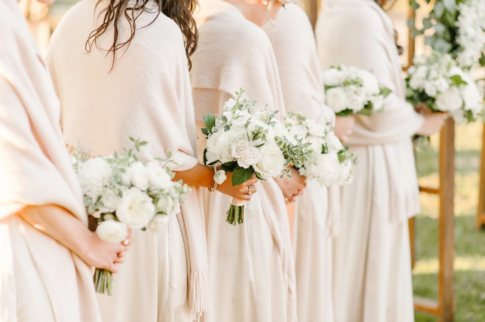 Row of bridesmaids in cream wraps/gowns holding all-white garden bouquets, backs to camera in golden light