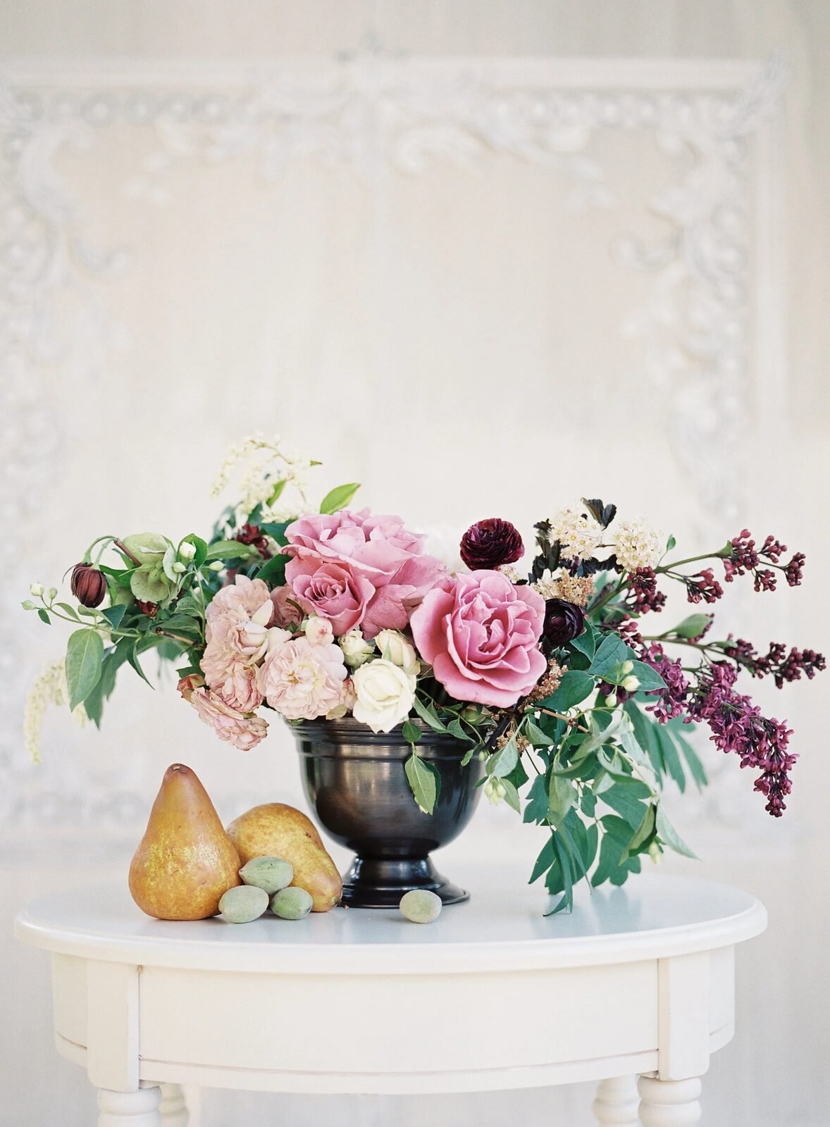 Fine-art film still life: pink/burgundy rose arrangement in dark urn against ornate plaster wall, pears at base