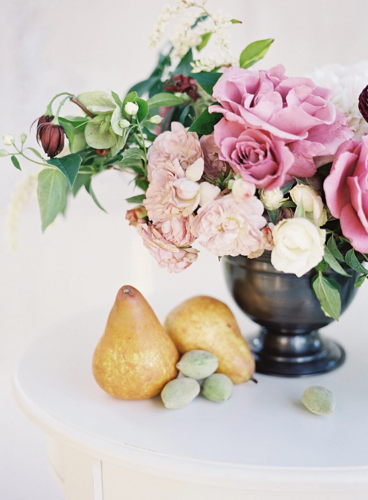 Fine-art film still life: mauve/blush rose arrangement in pewter urn with pears and almonds on cream table